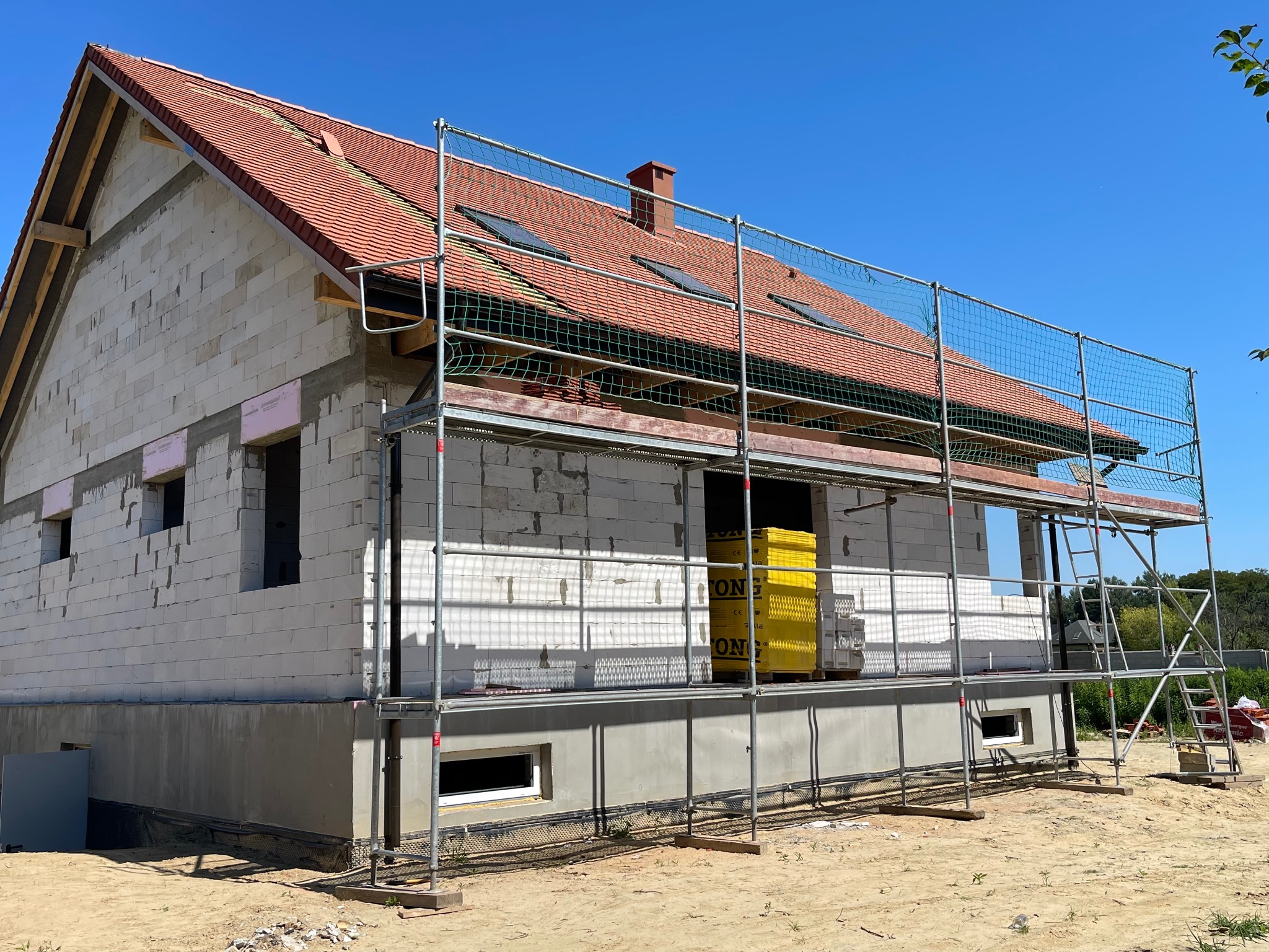 Dom w trakcie budowy z częściowo położonym czerwonym dachem, otoczony rusztowaniem z siatką zabezpieczającą. Widoczne okna dachowe i komin.