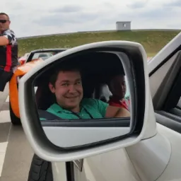 Uśmiechnięty mężczyzna w zielonej koszuli widoczny w lusterku bocznym białego samochodu, na torze wyścigowym, z innym samochodem w tle.