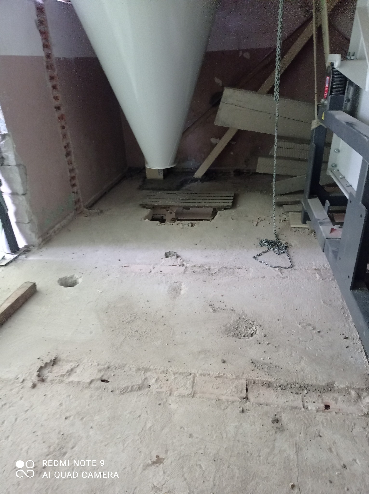 Wnętrze w trakcie remontu z widocznymi elementami konstrukcyjnymi, betonową podłogą z ubytkami, fragmentem białego lejka i łańcuchem zwisającym z sufitu.