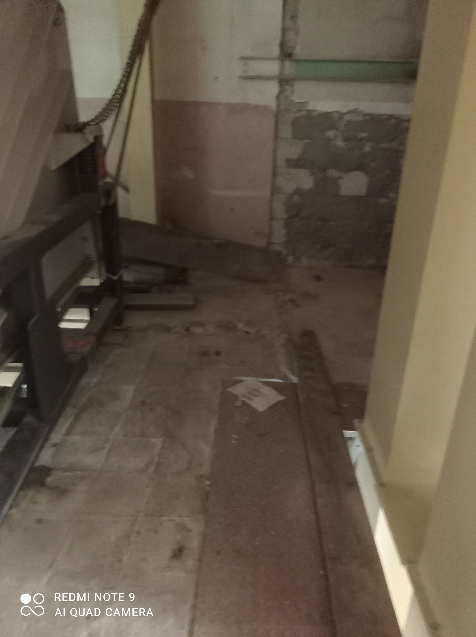 Wnętrze w trakcie remontu z widocznym fragmentem maszyny do obróbki materiałów, odsłoniętym murem i prowizorycznie zabezpieczoną podłogą.