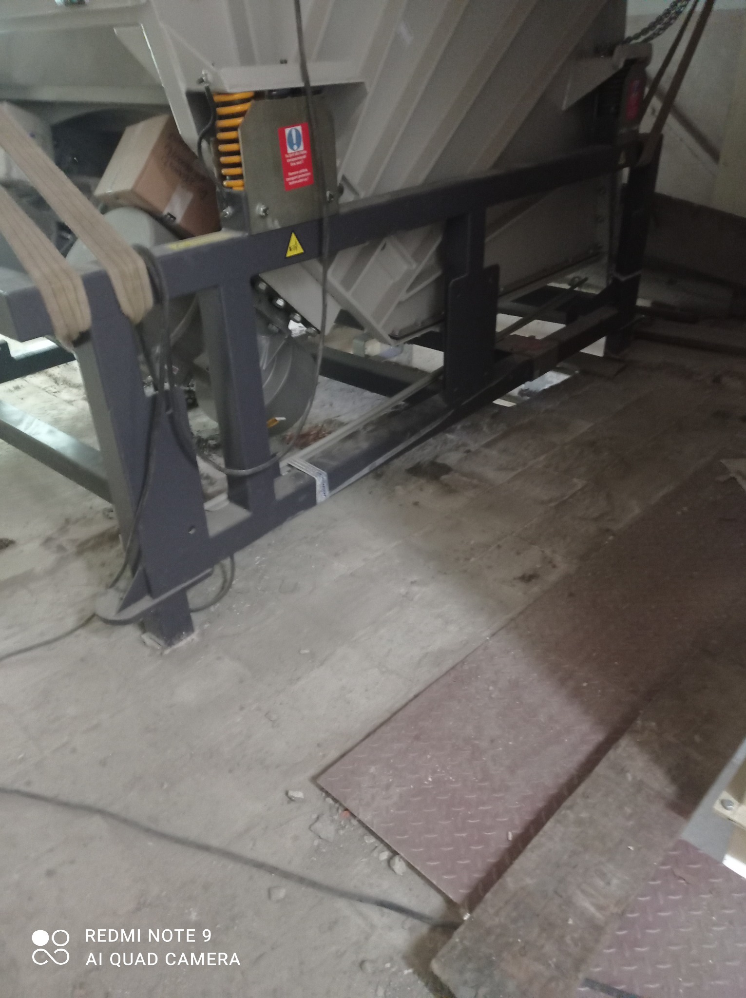 Fragment konstrukcji przemysłowej z szarym metalowym stelażem, widoczne elementy mechaniczne i ostrzegawcza naklejka z wykrzyknikiem na żółtym tle.