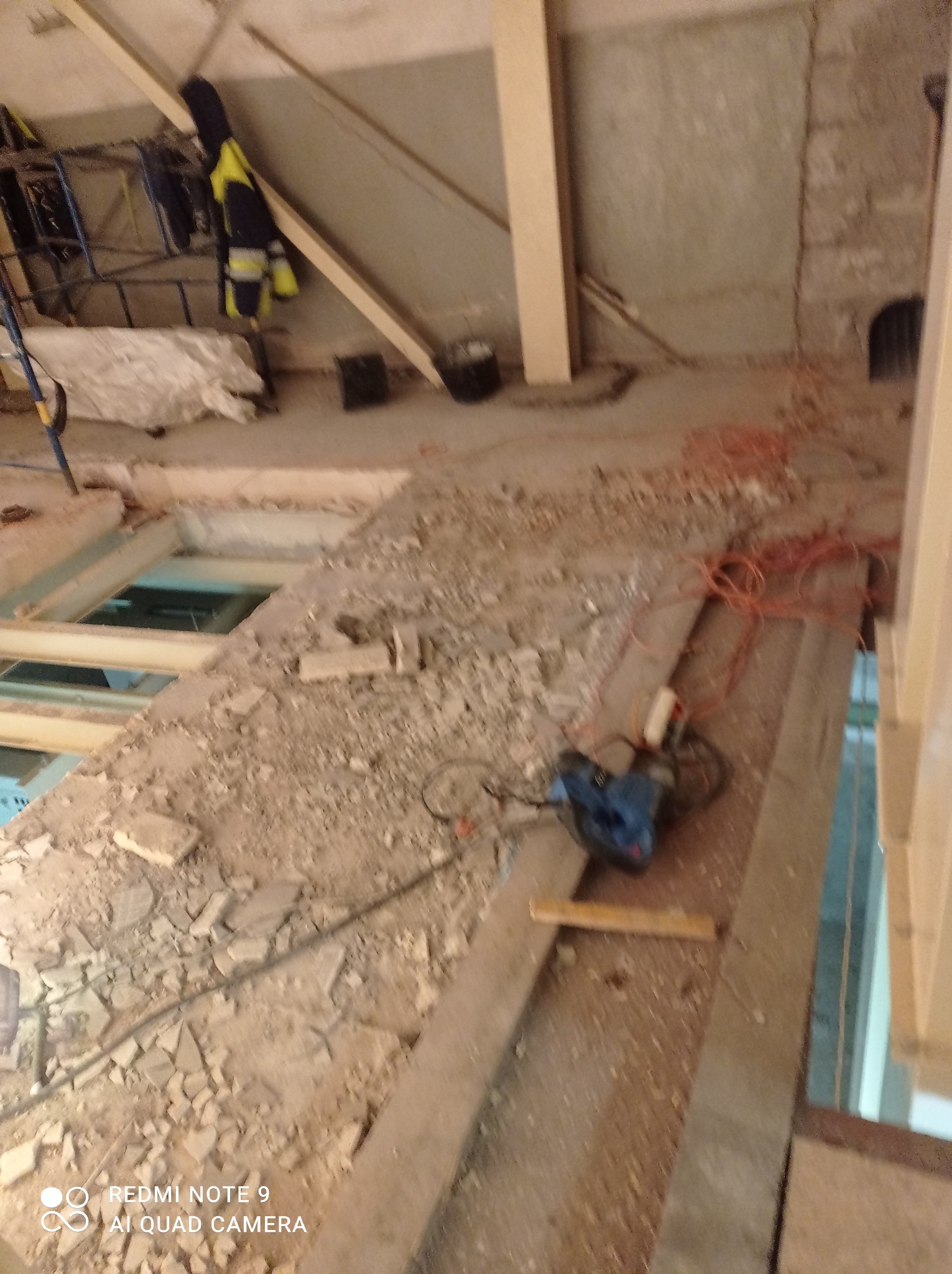 Demontaż podłogi na poddaszu: gruz, odsłonięte legary, widoczne narzędzie do cięcia, odzież robocza w tle.