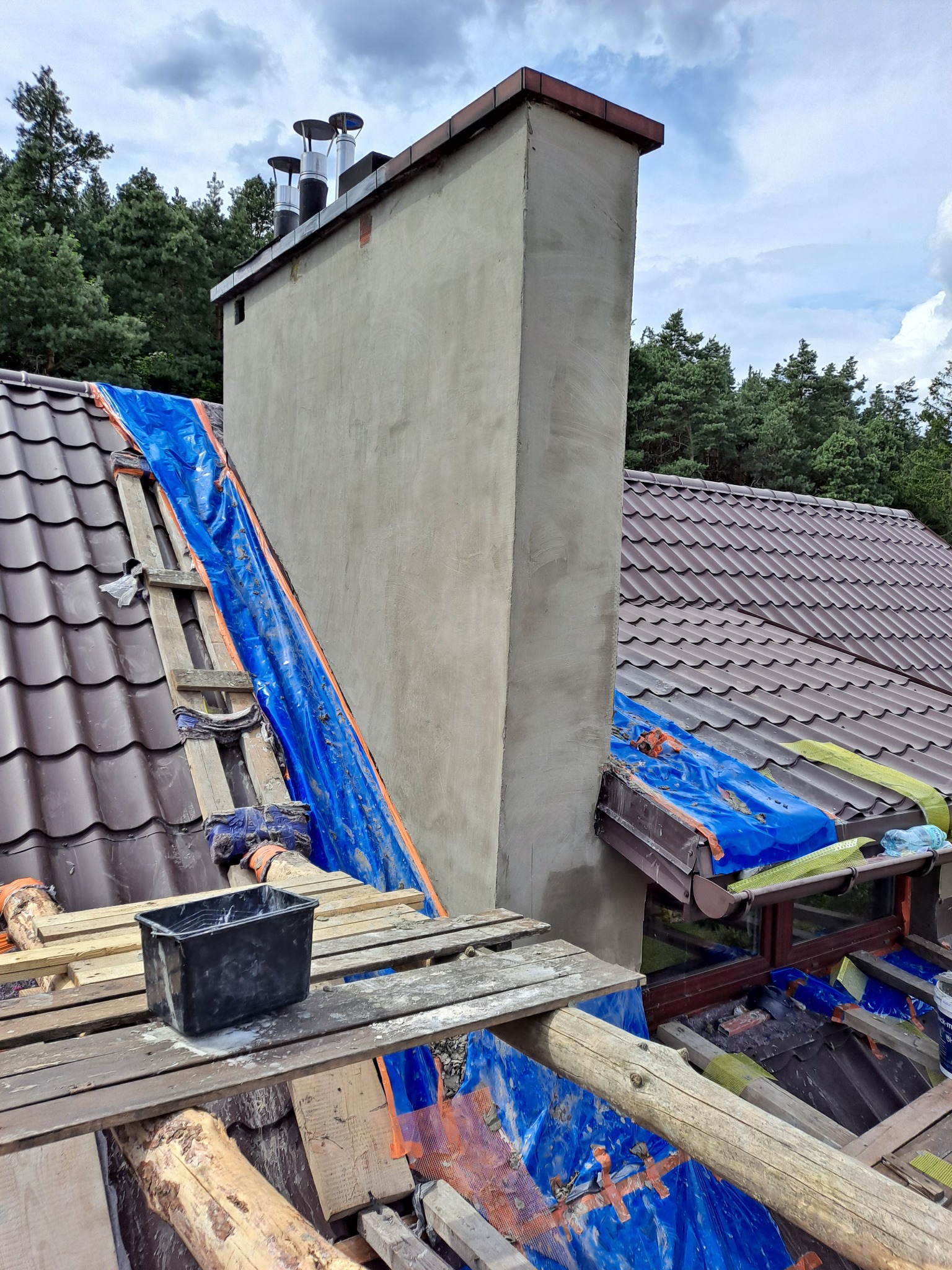Otynkowany komin na dachu z brązową dachówką, zabezpieczony niebieską folią i drewnianą konstrukcją, widok z góry na tle lasu i pochmurnego nieba.