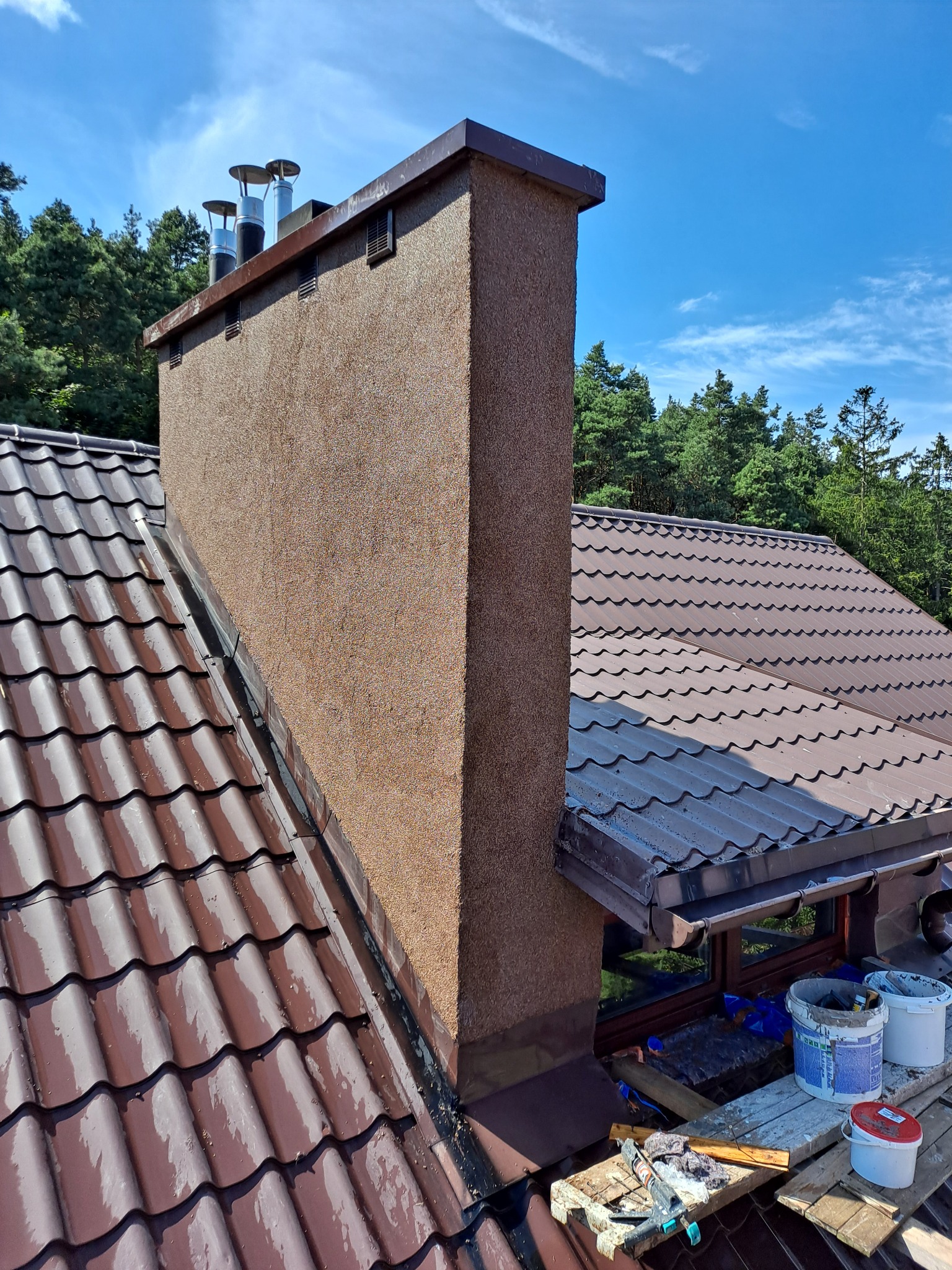 Widok z góry na brązowy dach z dachówki ceramicznej i komin z fakturą baranka, na dachu wiaderka z farbą i narzędzia, w tle las i błękitne niebo.