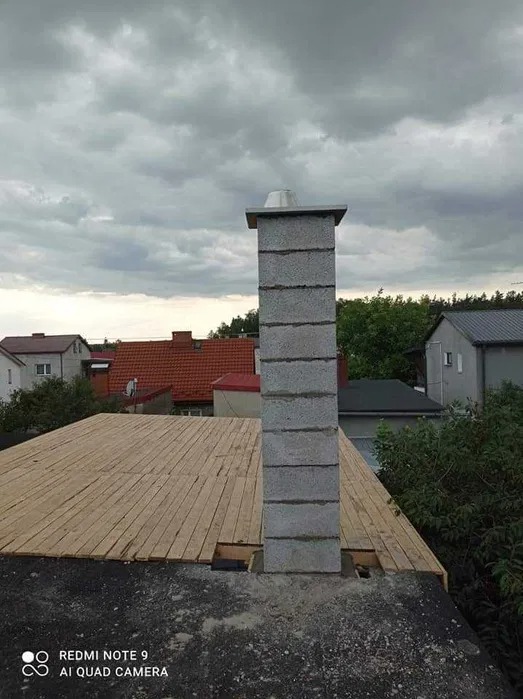 Wzniesiony komin z szarych bloczków na tle drewnianego poszycia dachu i pochmurnego nieba.