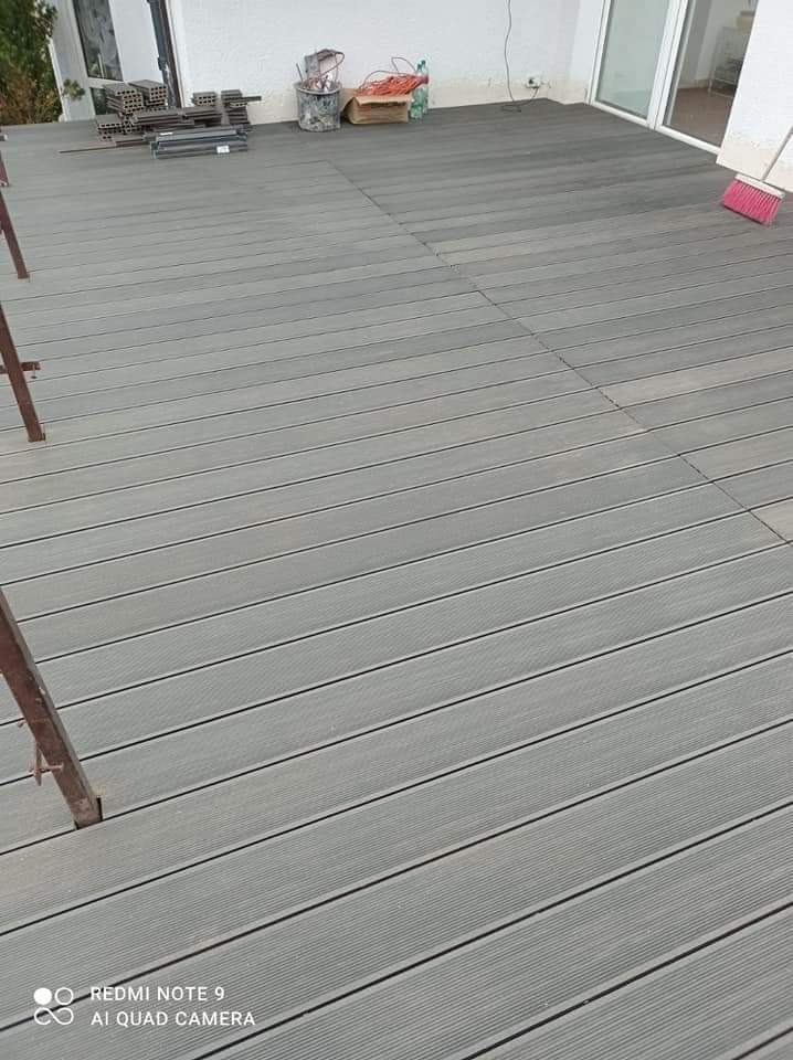 Szara deska tarasowa kompozytowa, częściowo ułożona, z widocznym fragmentem rusztowania, wiaderkiem z narzędziami i resztkami materiałów budowlanych w tle.