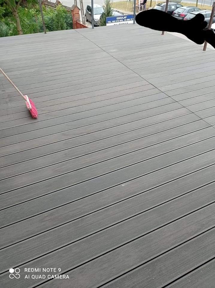 Szare panele podłogowe na balkonie, widoczna szczotka do zamiatania, w tle budynek z zaparkowanymi samochodami.