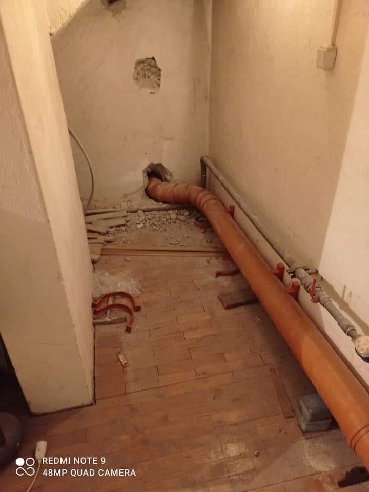 Wnętrze w trakcie remontu łazienki: brązowa rura kanalizacyjna w ścianie, widoczne ślady po kuciu i starej instalacji, drewniana podłoga, surowy stan wykończenia.