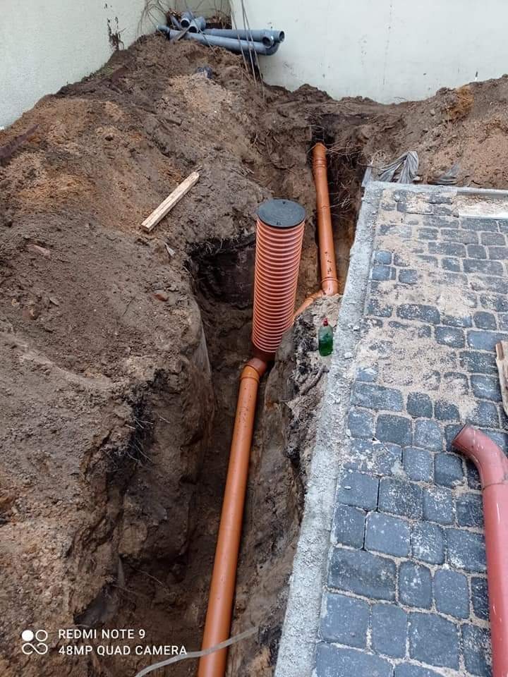 Wykop z ułożonymi rurami drenarskimi i studzienką rewizyjną, obok ułożona kostka brukowa, widoczne ślady ziemi na kostce.