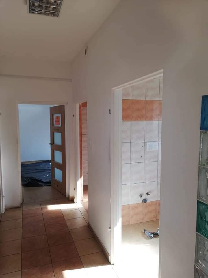 Korytarz w trakcie remontu z widoczną łazienką z ułożonymi płytkami w odcieniach bieli i pomarańczy, narzędzia na podłodze, widoczne płytki podłogowe w korytarzu i drzwi z szybami w tle.