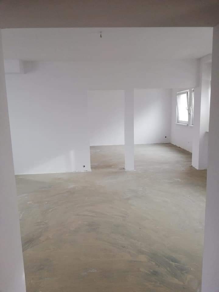 Surowe wnętrze w trakcie przygotowania do prac wykończeniowych, widoczne surowe ściany i podłoga, z kolumnami podtrzymującymi strop, okno z zasłoniętą roletą.