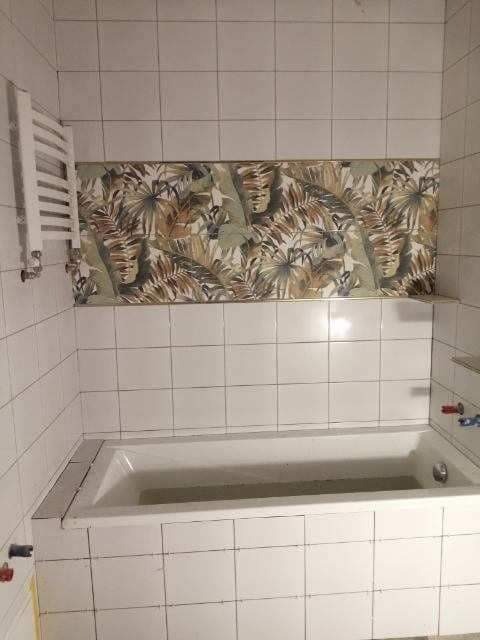 Biała wanna obłożona białymi, kwadratowymi płytkami, nad wanną pas płytek z motywem roślinnym, po lewej stronie grzejnik drabinkowy.