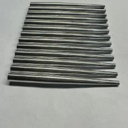 Rząd równolegle ułożonych, precyzyjnie obrobionych, stożkowych prętów ze stali nierdzewnej, o gładkiej, lustrzanej powierzchni, leżących na szarym tle.