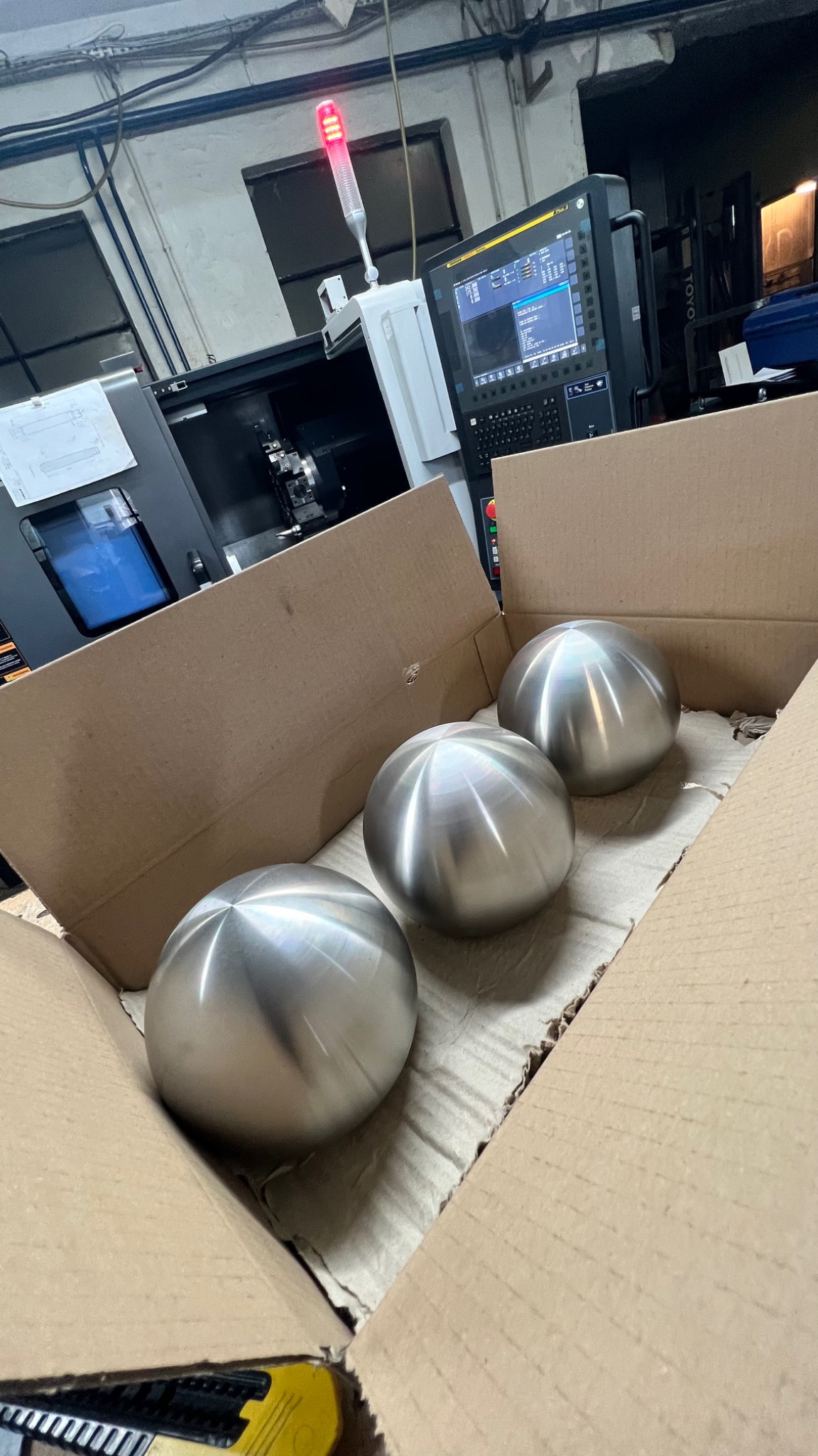 Trzy metalowe kule w kartonowym pudełku, w tle widoczna maszyna CNC z czerwonym światłem sygnalizacyjnym.