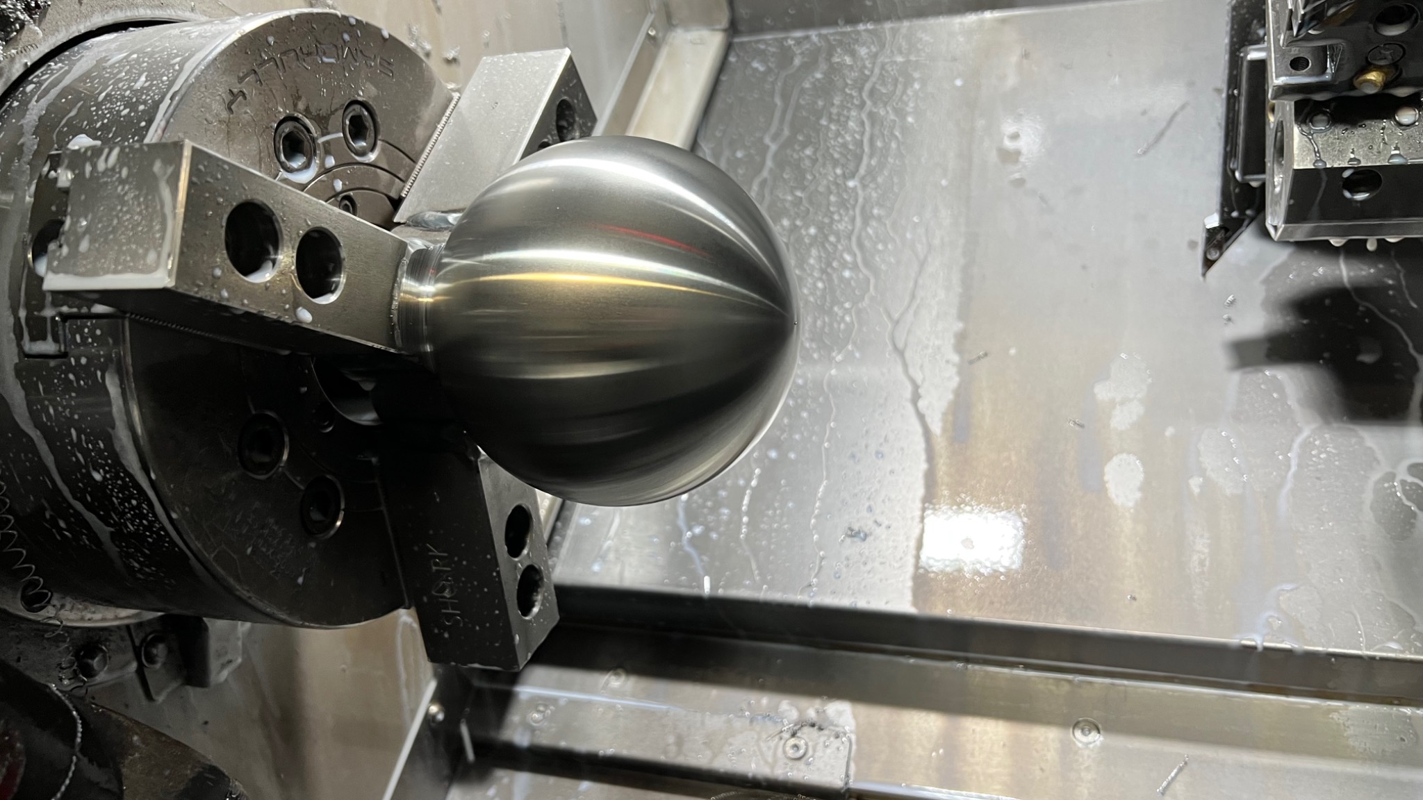 Metalowa kula obrabiana w uchwycie tokarskim CNC, widoczne ślady chłodziwa i obróbki.