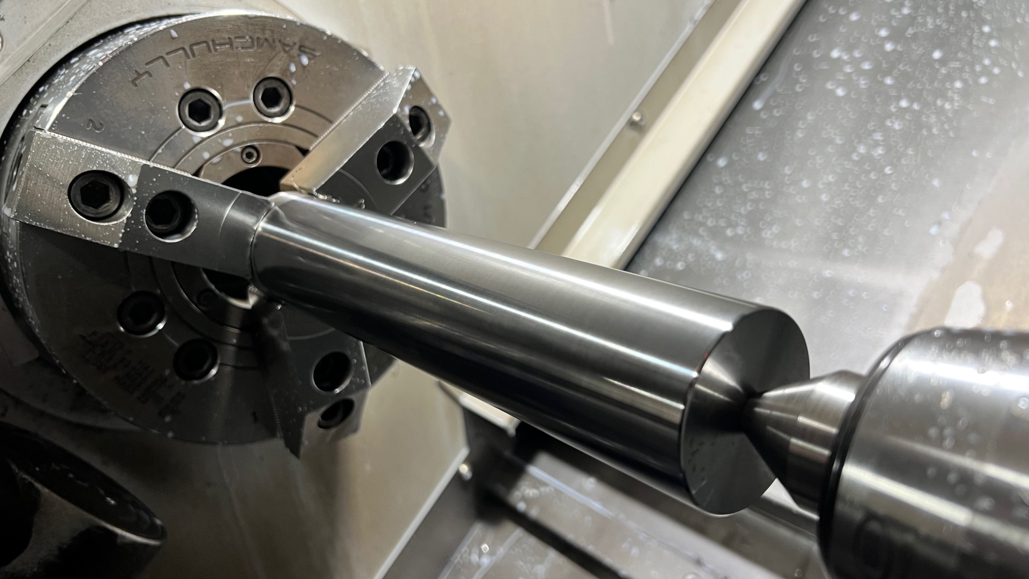 Obrabiany metalowy element o kształcie stożka w uchwycie tokarskim podczas pracy, widoczne chłodziwo i wióry.