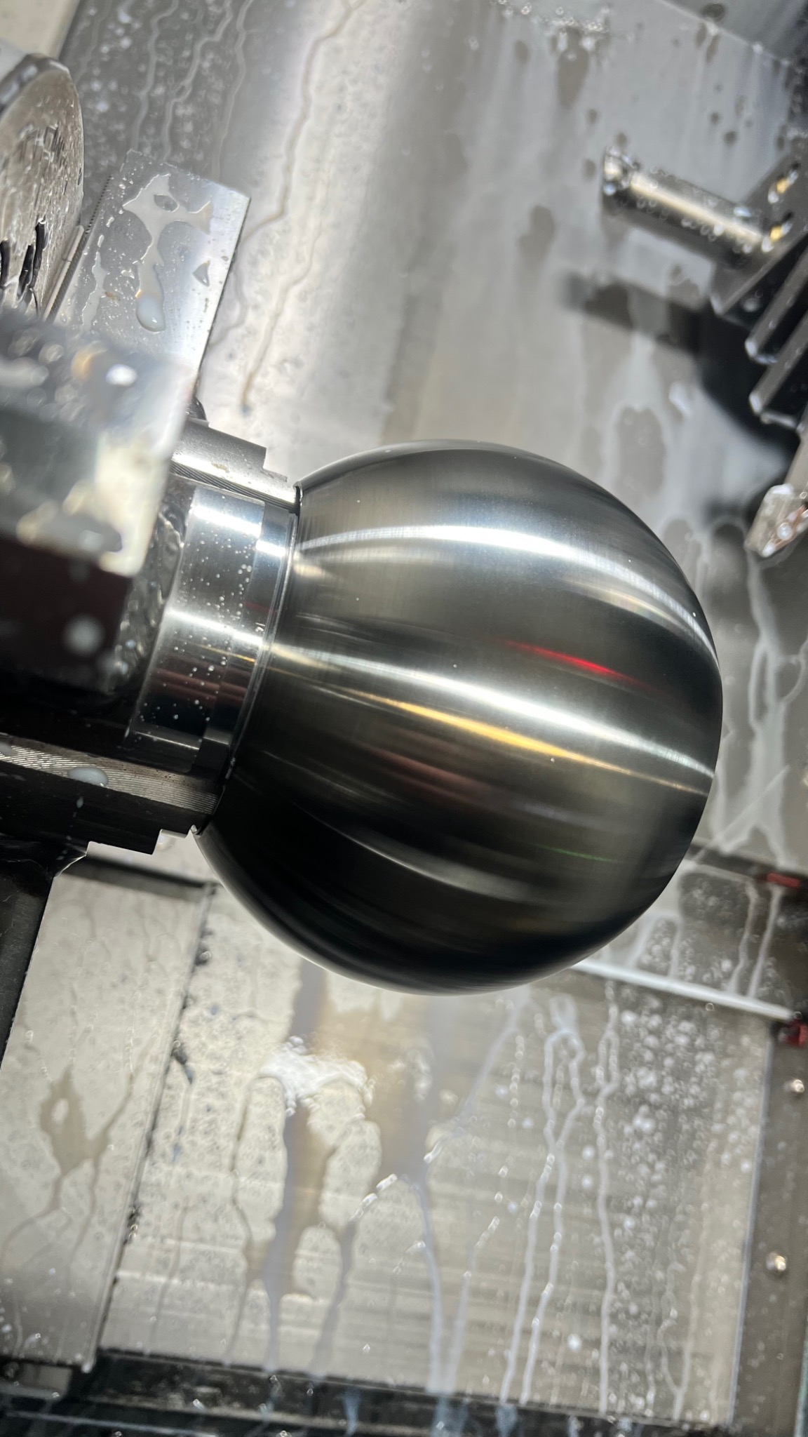 Obróbka metalowej kuli na tokarce CNC, widoczne chłodziwo i narzędzie skrawające w trakcie pracy.