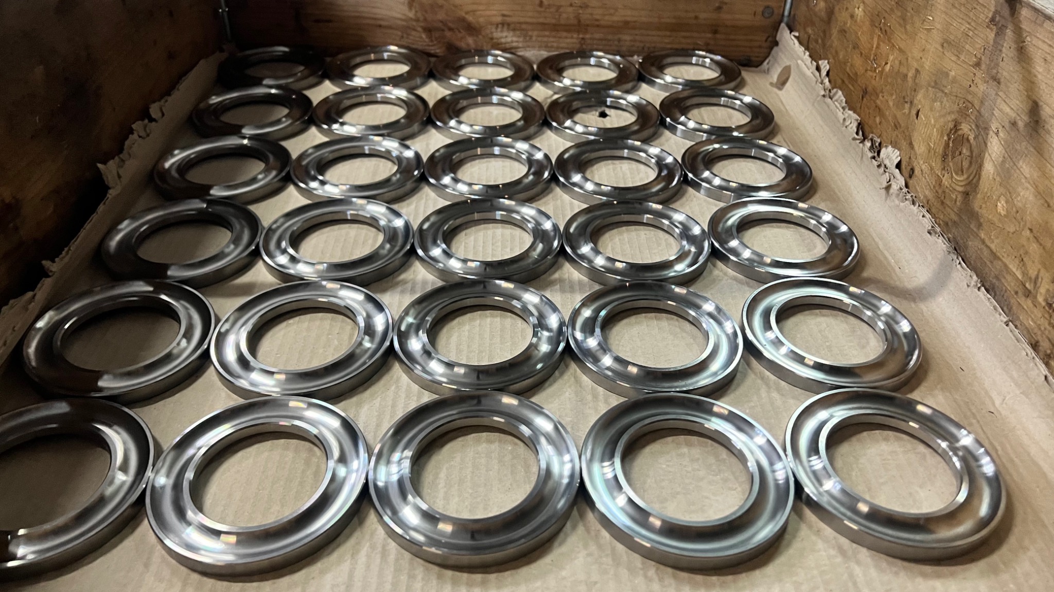 Partia metalowych pierścieni z kołnierzem, ułożonych rzędami w drewnianej skrzyni wyłożonej papierem, widoczne refleksy światła na gładkiej powierzchni.
