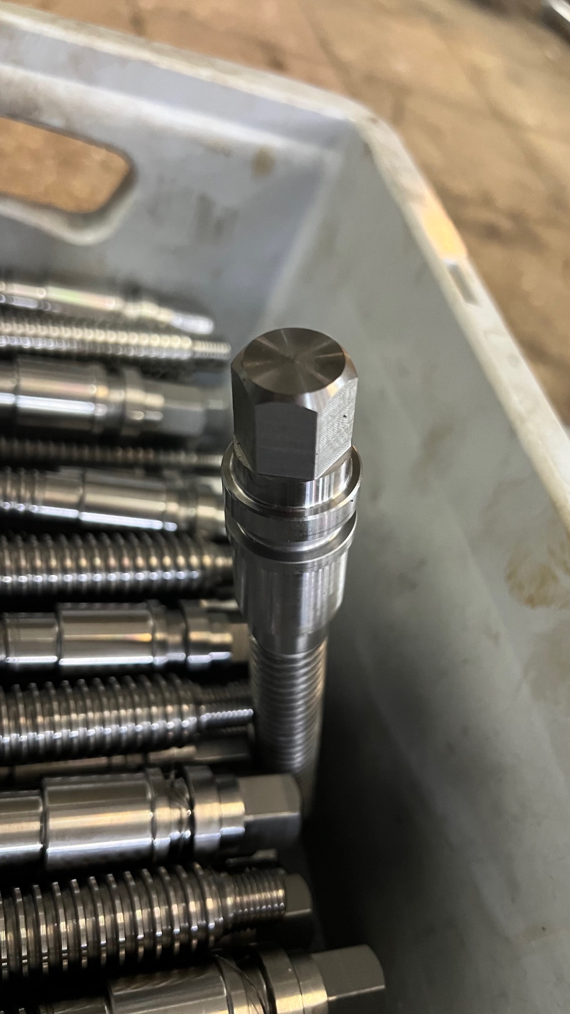 Metalowe elementy z gwintem i sześciokątną główką ułożone w plastikowym pojemniku, jeden element wystaje pionowo do góry, widoczne ślady obróbki skrawaniem.