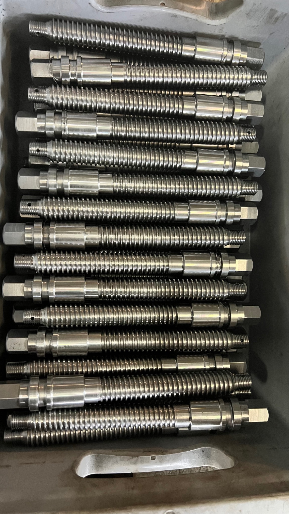 Partie precyzyjnie obrobionych, metalowych elementów z gwintem i sześciokątnym zakończeniem, ułożonych równolegle w metalowym pojemniku.