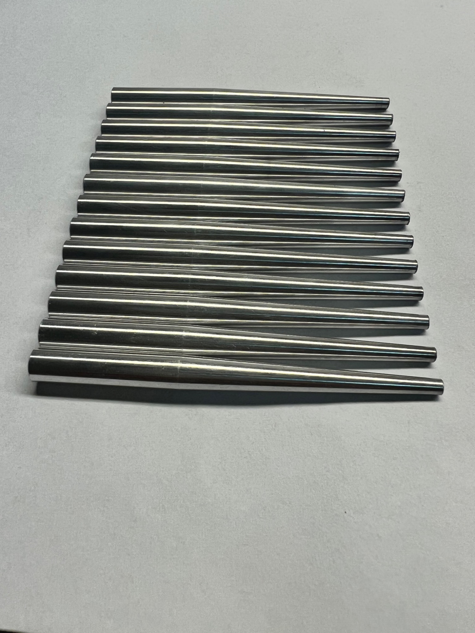 Rząd równolegle ułożonych, precyzyjnie obrobionych, stożkowych prętów ze stali nierdzewnej, o gładkiej, lustrzanej powierzchni, leżących na szarym tle.