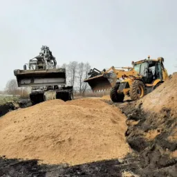 Dwie maszyny budowlane, koparka i ładowarka, rozładowują piasek do wykopu w pochmurny dzień. W tle widoczne drzewa.