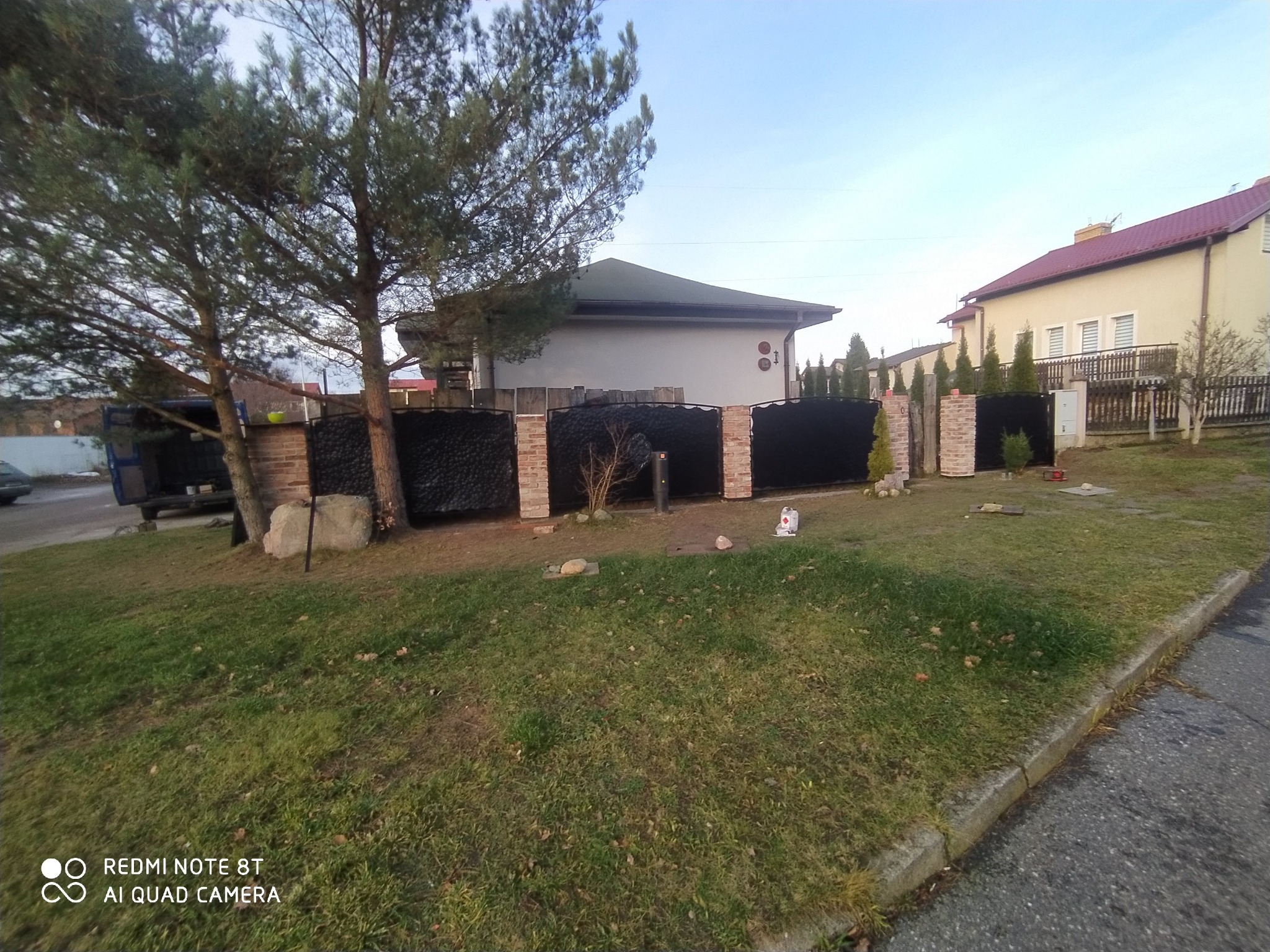 Nowoczesne ogrodzenie panelowe z elementami murowanymi z cegły klinkierowej, widoczne z zewnątrz posesji, z fragmentem trawnika i drzew.