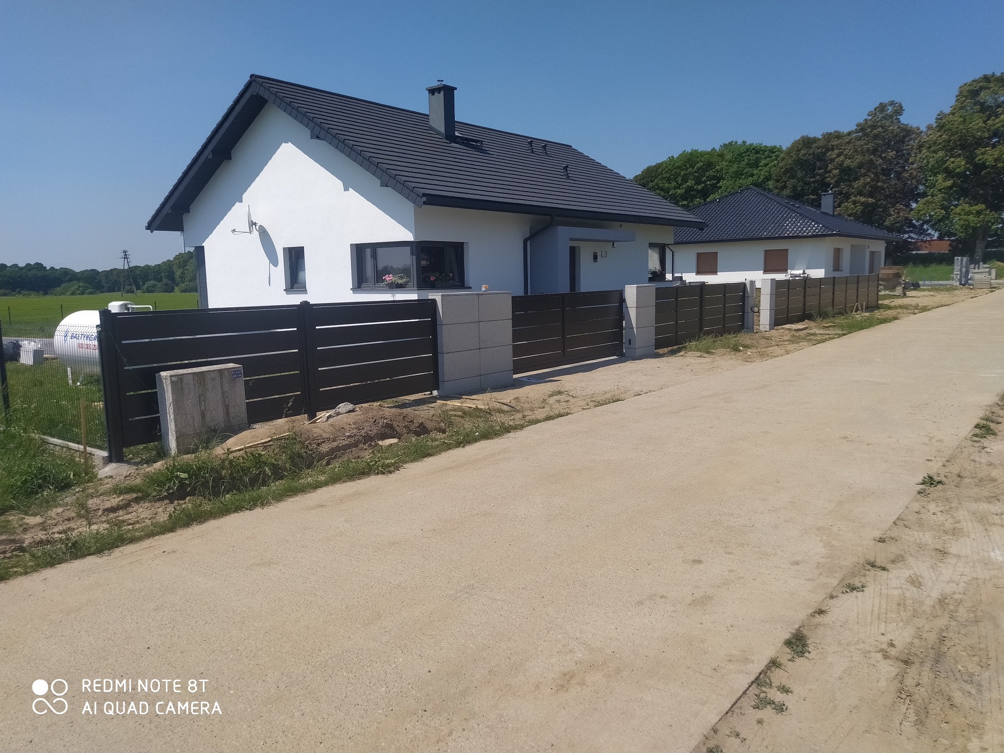 Nowoczesne ogrodzenie panelowe w kolorze antracytowym, oddzielające posesję z białym domem o ciemnej dachówce, widok z perspektywy drogi gruntowej w słoneczny dzień.