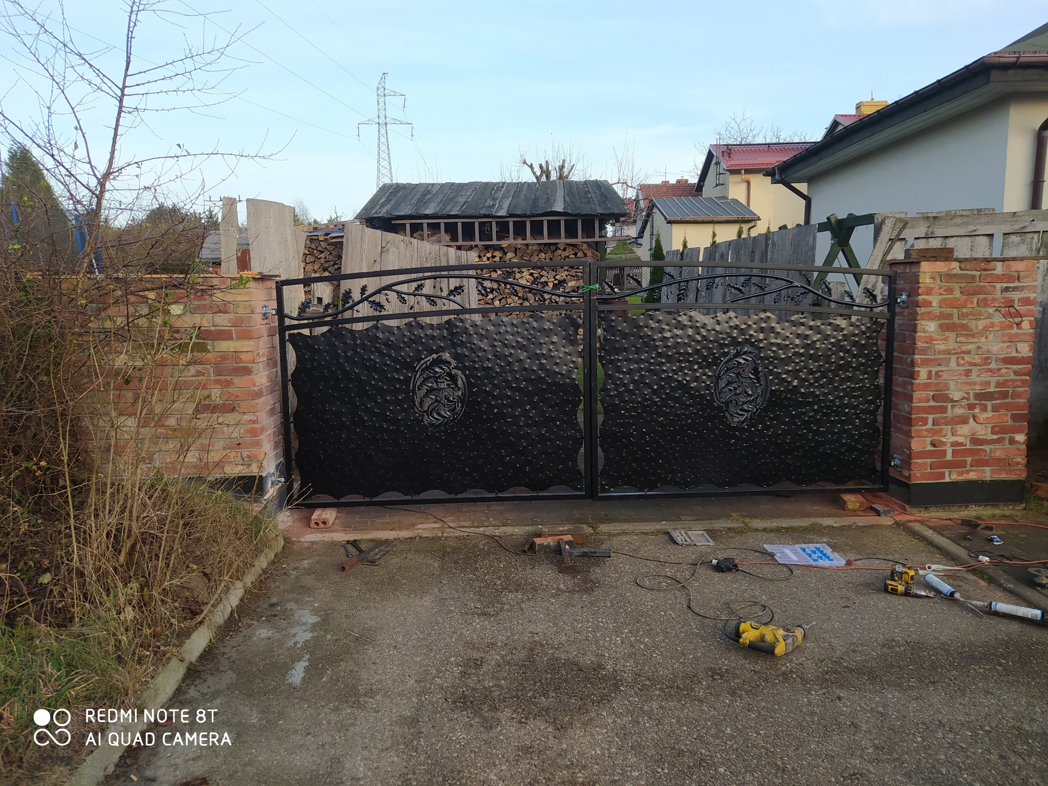 Czarna, dwuskrzydłowa brama wjazdowa z ozdobnymi elementami, zamontowana na ceglanych słupkach, widoczne narzędzia i akcesoria montażowe na betonowym podjeździe.