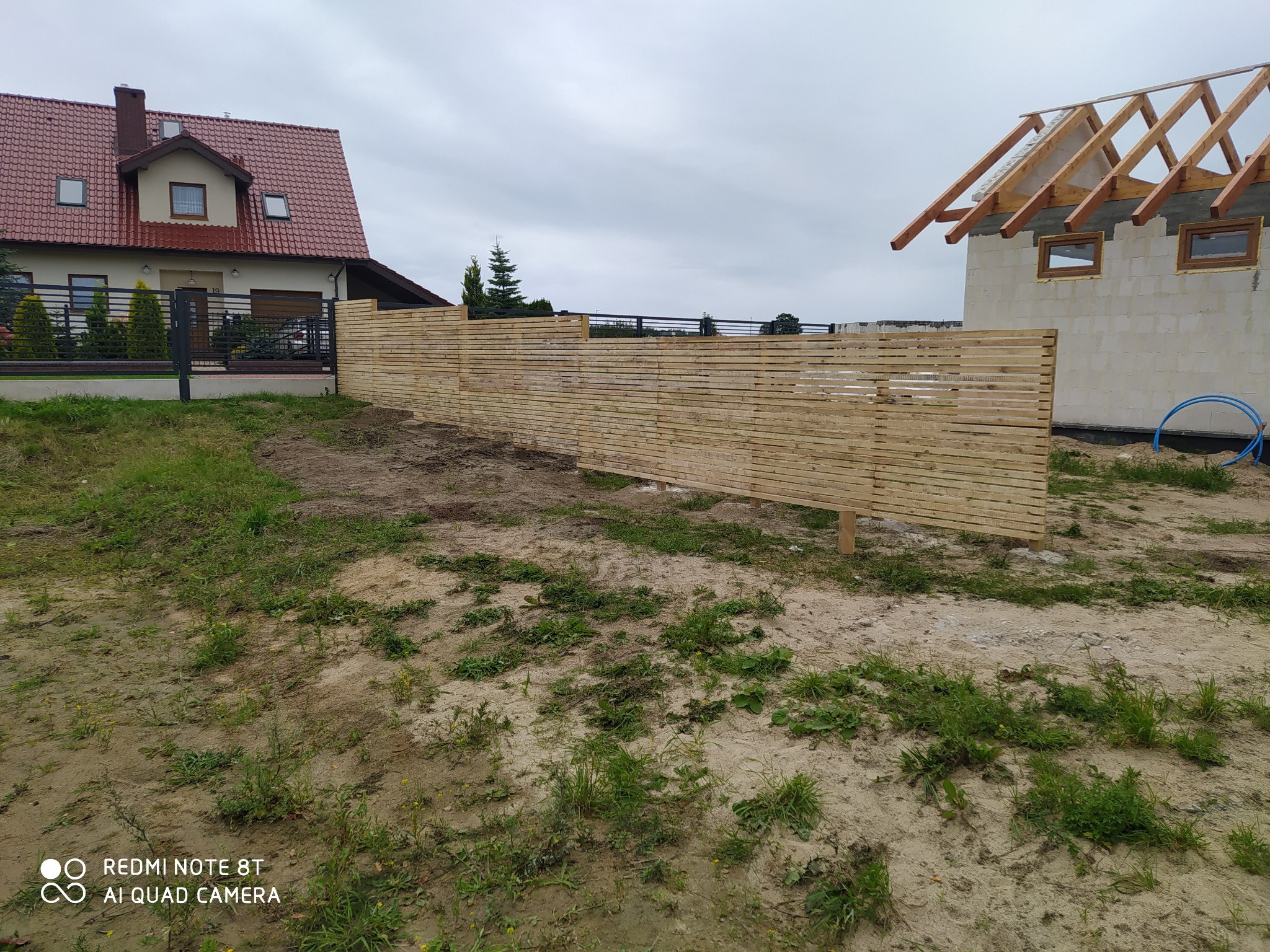 Nowoczesne ogrodzenie panelowe z jasnego drewna, widoczne na tle domu z czerwoną dachówką i budowy w stanie surowym otwartym, pochmurne niebo.