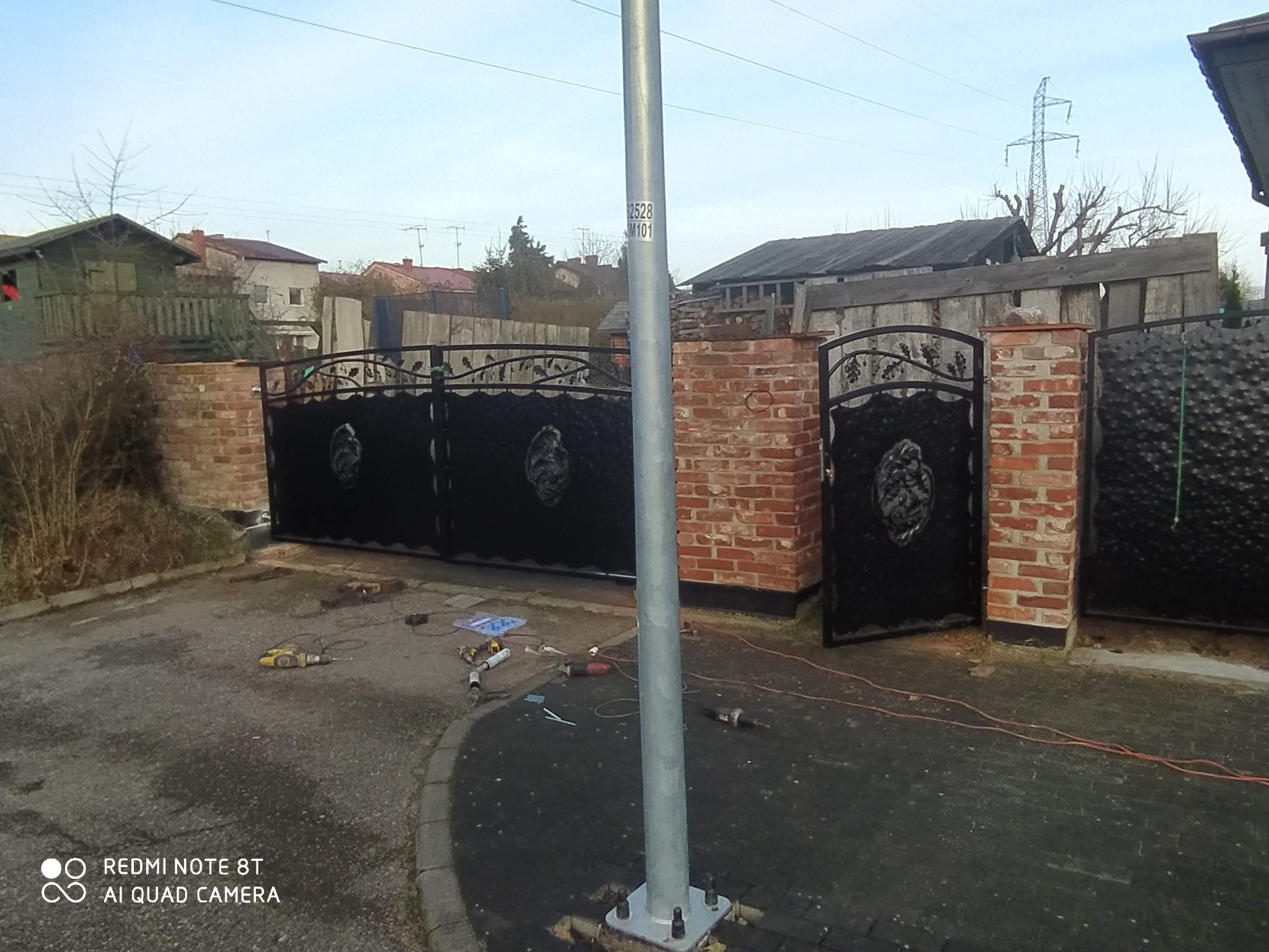 Nowo zamontowana, czarna, dwuskrzydłowa brama z dekoracyjnymi elementami, osadzona na ceglanych słupkach, widoczne narzędzia montażowe na podłożu.