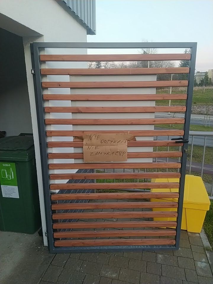 Nowoczesna brama z poziomych drewnianych listew w szarej metalowej ramie, z kartką 'NIE DOTYKAĆ! NIE ZAMYKAĆ!?', obok zielony i żółty pojemnik na śmieci.