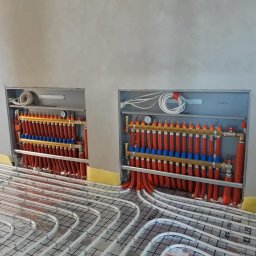 Hydraulik w mieście Dominik Wydmuch - Rozdzielacze ogrzewania podłogowego wnękowe z rurami grzewczymi ułożonymi na siatce, widok z góry, szare ściany w tle, żółta taśma brzegowa.