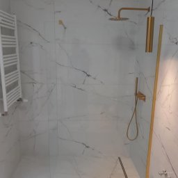 Hydraulik w mieście Dominik Wydmuch - Nowoczesna kabina prysznicowa walk-in z białymi płytkami imitującymi marmur, złotą armaturą i odpływem liniowym. Minimalistyczny design z grzejnikiem drabinkowym.