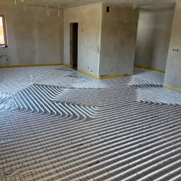 Hydraulik w mieście Dominik Wydmuch - Instalacja ogrzewania podłogowego w Częstochowie. Białe rurki ułożone na siatce w surowym wnętrzu, przygotowane do zalania wylewką. Widoczne okno i surowe ściany.