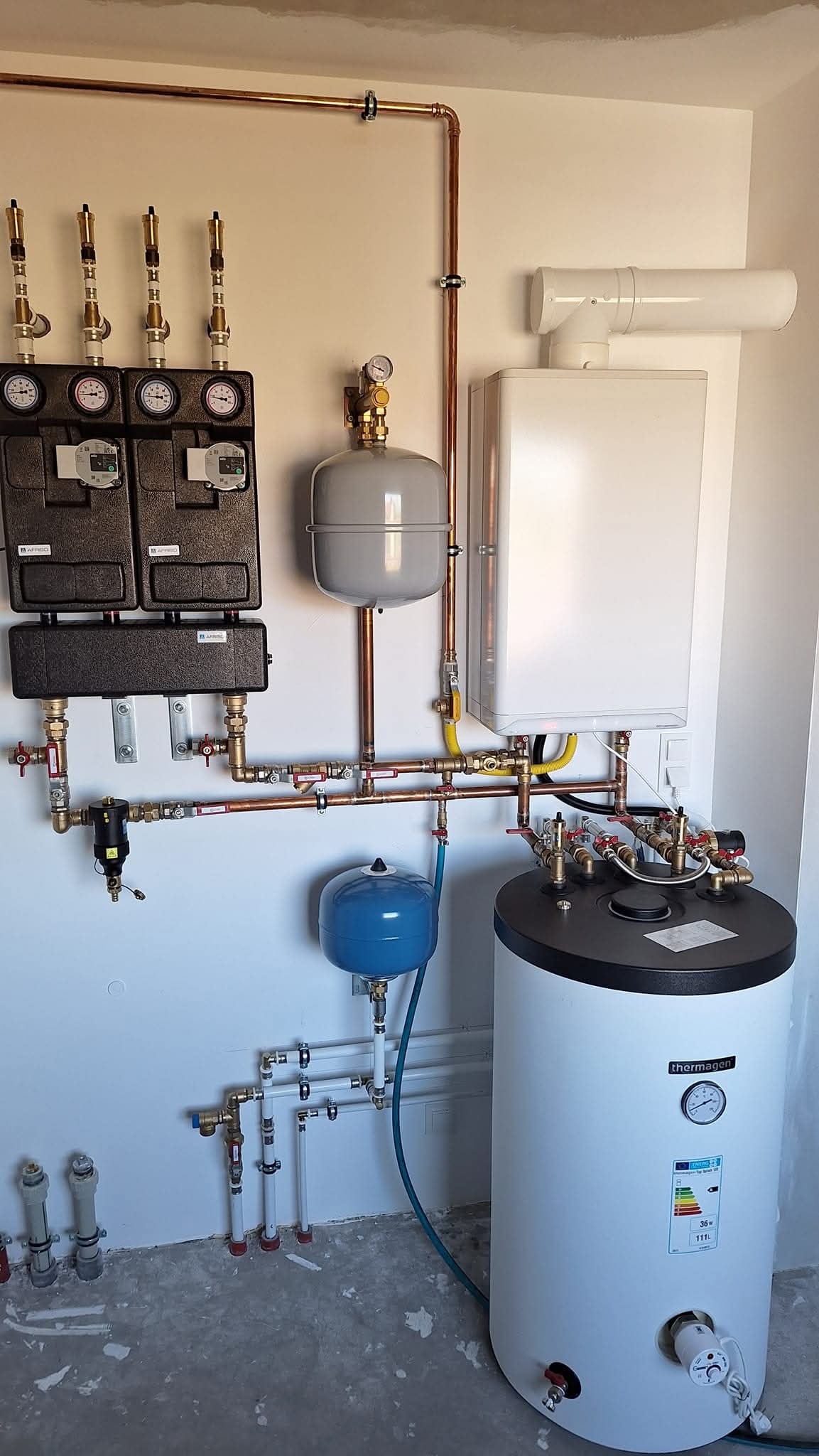 Kompleksowa instalacja grzewcza z piecem Thermagen, zbiornikiem, rozdzielaczami Afriso i miedzianymi rurami, zamontowana na ścianie w surowym wnętrzu.