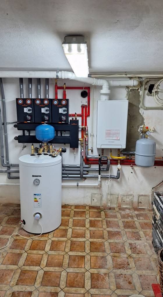 Złożona instalacja grzewcza w piwnicy: biały bojler, piec, liczne rury miedziane i szare, czarne panele sterujące z manometrami, podświetlone jarzeniówką.