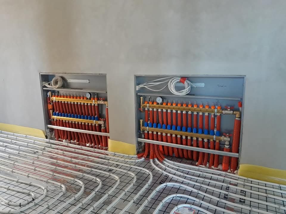 Rozdzielacze ogrzewania podłogowego wnękowe z rurami grzewczymi ułożonymi na siatce, widok z góry, szare ściany w tle, żółta taśma brzegowa.