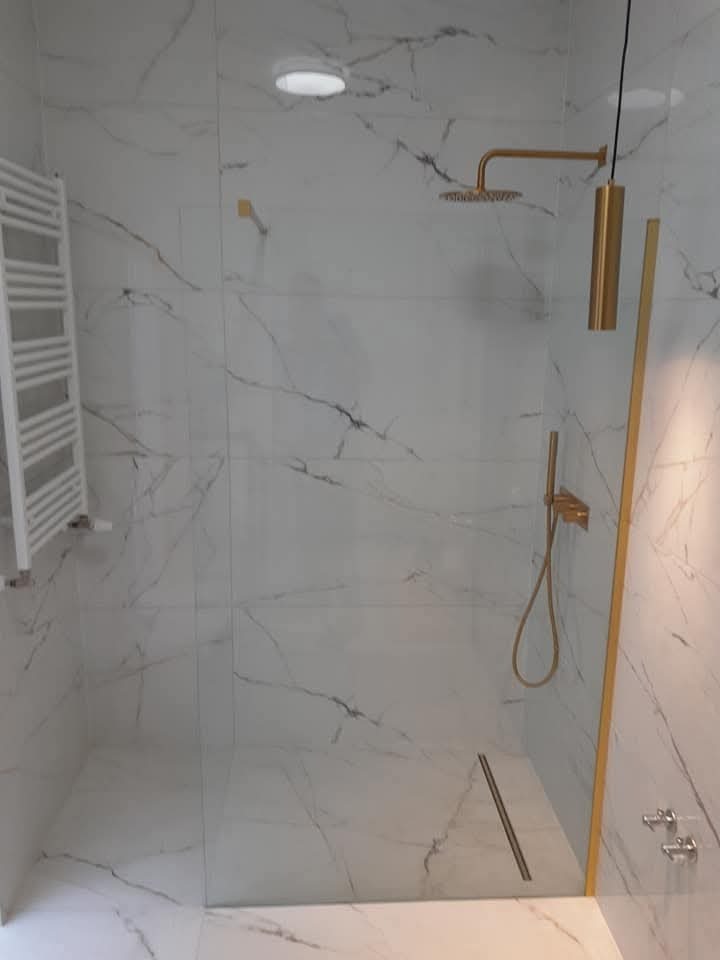 Nowoczesna kabina prysznicowa walk-in z białymi płytkami imitującymi marmur, złotą armaturą i odpływem liniowym. Minimalistyczny design z grzejnikiem drabinkowym.