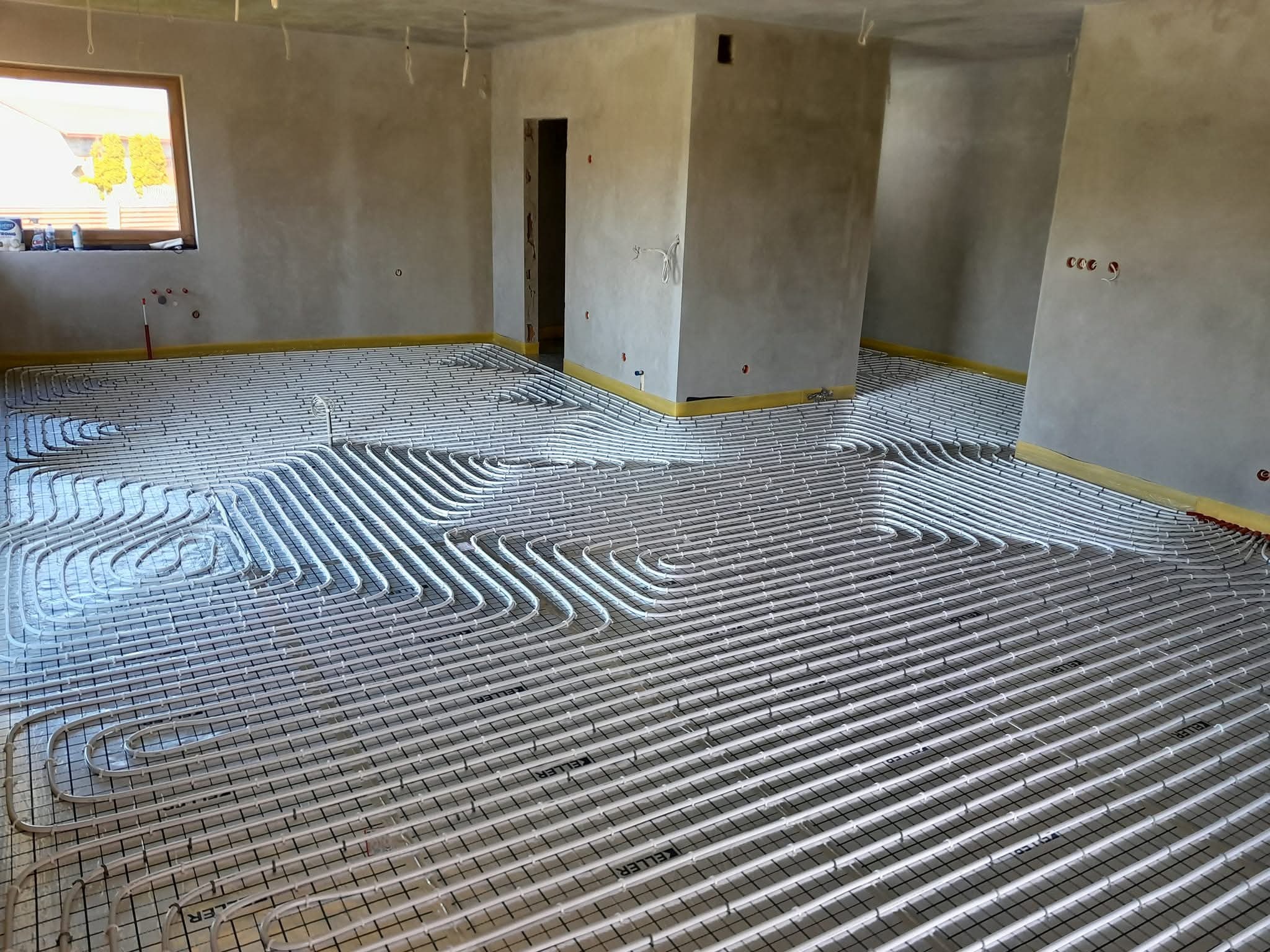 Instalacja ogrzewania podłogowego w Częstochowie. Białe rurki ułożone na siatce w surowym wnętrzu, przygotowane do zalania wylewką. Widoczne okno i surowe ściany.