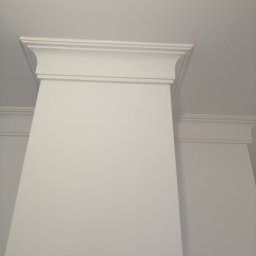 Sławomir Przybylski - Biała ściana z dekoracyjnym, sztukateryjnym wykończeniem pod sufitem, widok z dołu, z delikatnym cieniem.