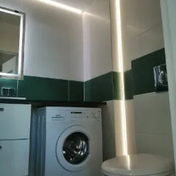 Nowoczesna łazienka z białymi i zielonymi płytkami, podświetlanym lustrem, pralką MPM i podtynkową spłuczką WC.