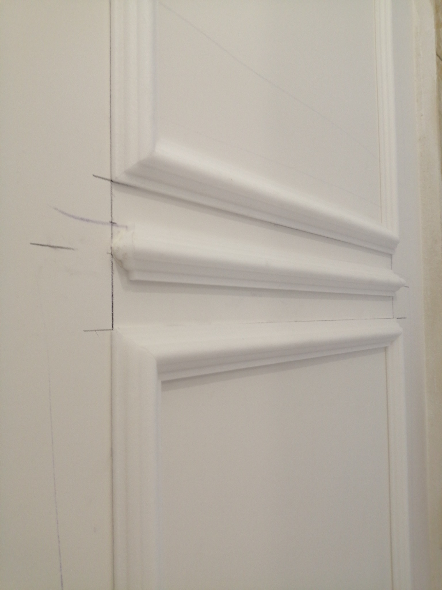 Fragment białej ściany z dekoracyjnymi listwami, zaznaczone ołówkiem linie pomocnicze przed montażem, widoczne niedoskonałości wykończenia.