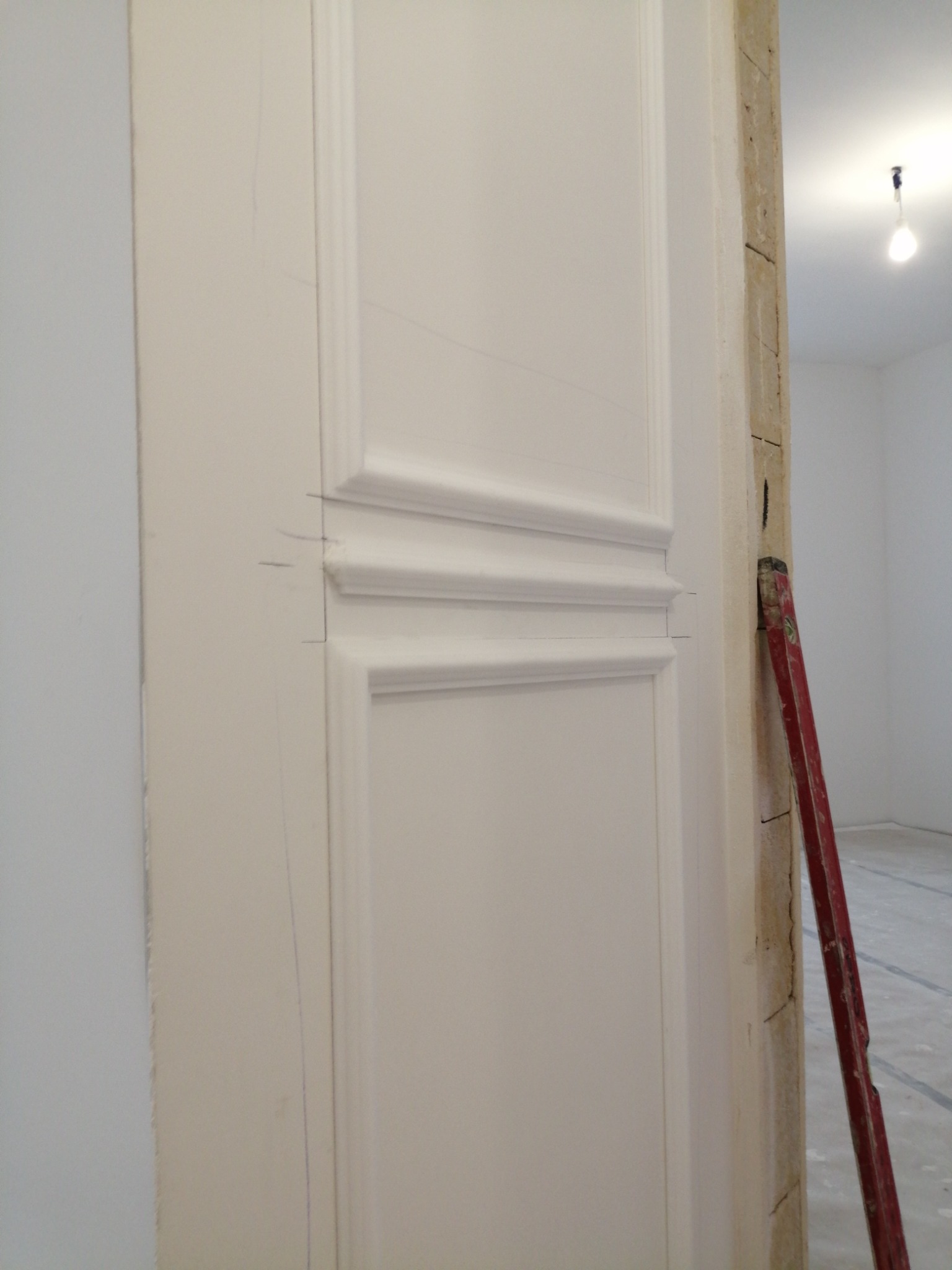 Fragment ściany z zamontowaną białą listwą przypodłogową z zaznaczonymi liniami cięcia, obok czerwona poziomica oparta o surową ścianę, w tle widoczne puste pomieszczenie z lampą na suficie.