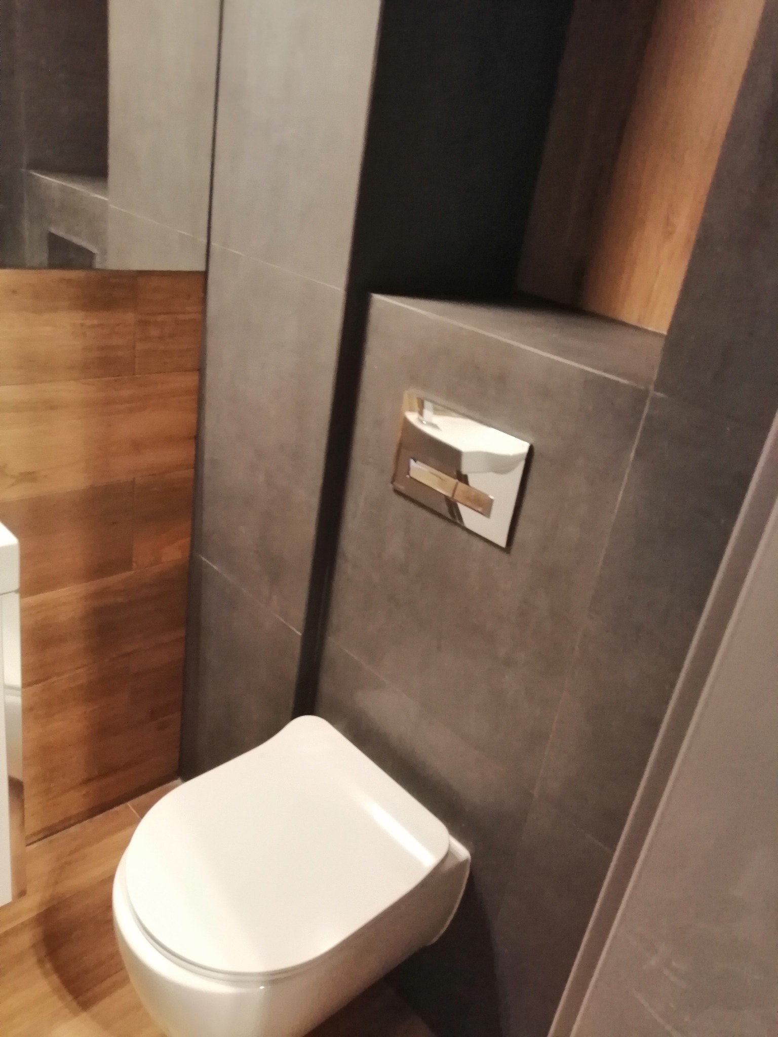 Nowoczesna toaleta podwieszana z chromowanym przyciskiem spłukującym na szarej, płytkowej ścianie, uzupełniona drewnopodobną okładziną.