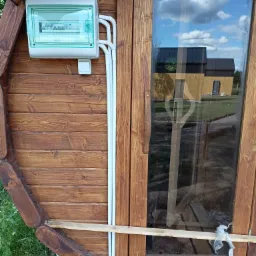 Skrzynka elektryczna natynkowa z zielonymi akcentami, zamontowana na drewnianej ścianie domku, z widocznymi białymi rurkami instalacyjnymi i odbiciem nieba w oknie.