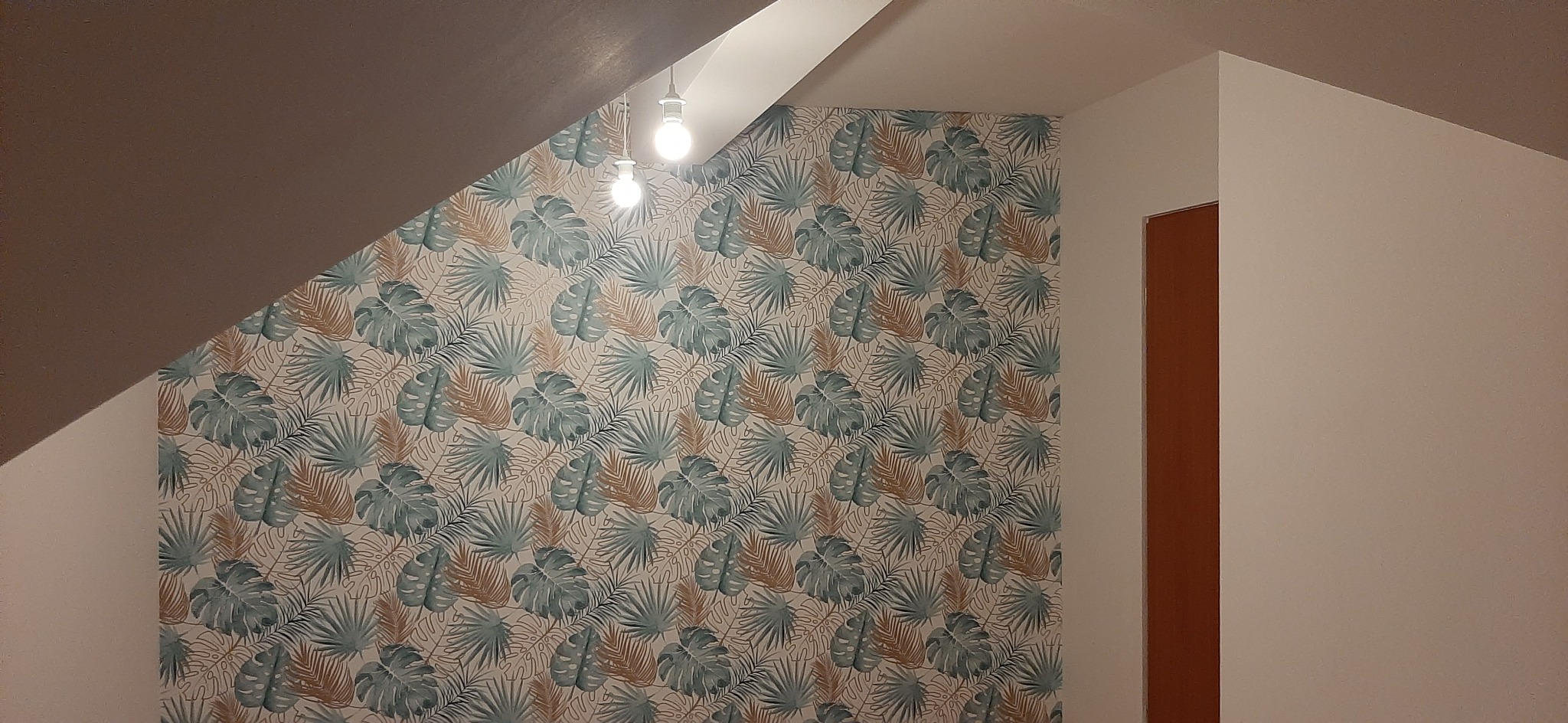 Ściana w pokoju na poddaszu wykończona tapetą w tropikalne liście, widoczne dwie żarówki i fragment brązowych drzwi.
