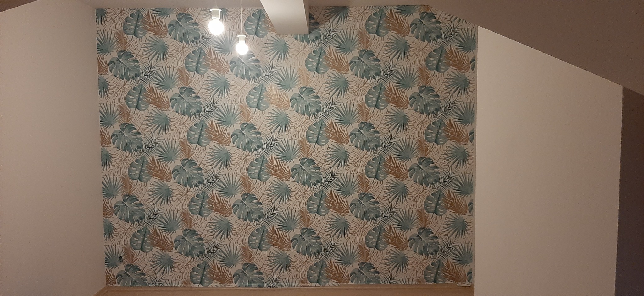 Ściana pokryta tapetą z motywem tropikalnych liści w odcieniach zieleni i brązu, oświetlona trzema żarówkami na suficie pod skosem.