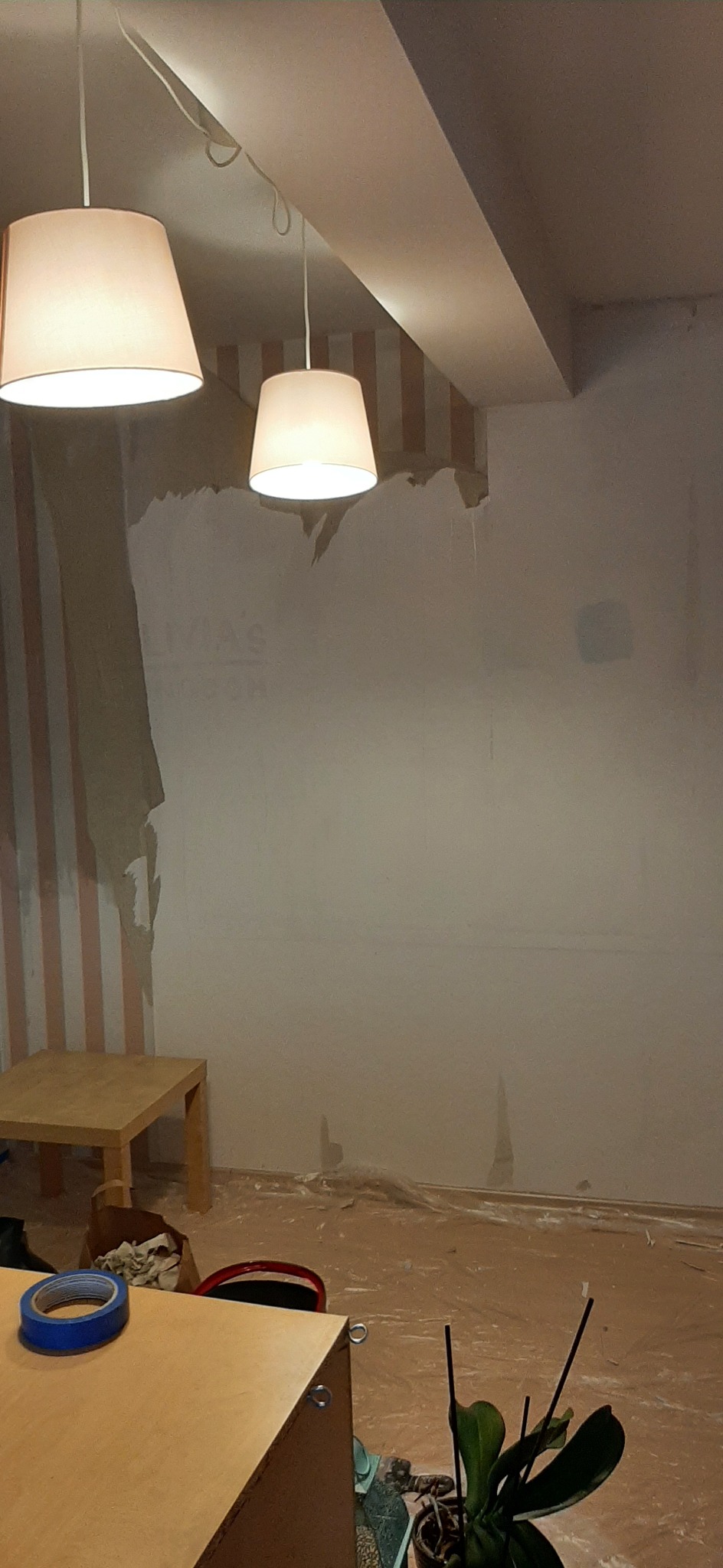 Pokój w trakcie przygotowania do prac wykończeniowych: resztki tapety, zagruntowana ściana, zabezpieczona podłoga, widoczne lampy i meble.