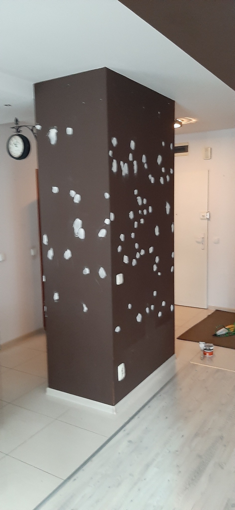 Brązowy filar z białymi plamami szpachli w jasnym wnętrzu, widoczny fragment podłogi z płytek, zegar na ścianie w tle.